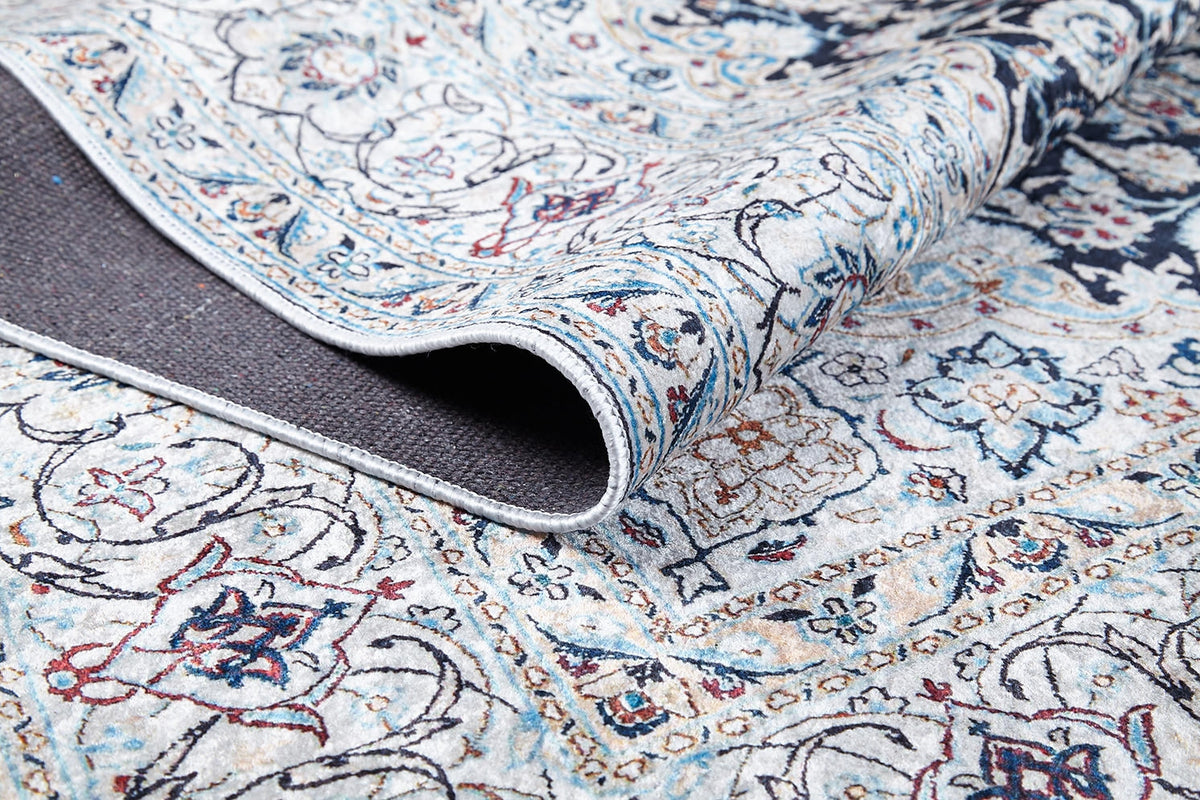 A close-up of the Antique Enchantment - Washable Rug - JR1686 shows intricate blue, red, and beige flat-weave patterns with a folded corner revealing its dark backing. Floral and geometric designs offer an allergy-friendly touch.