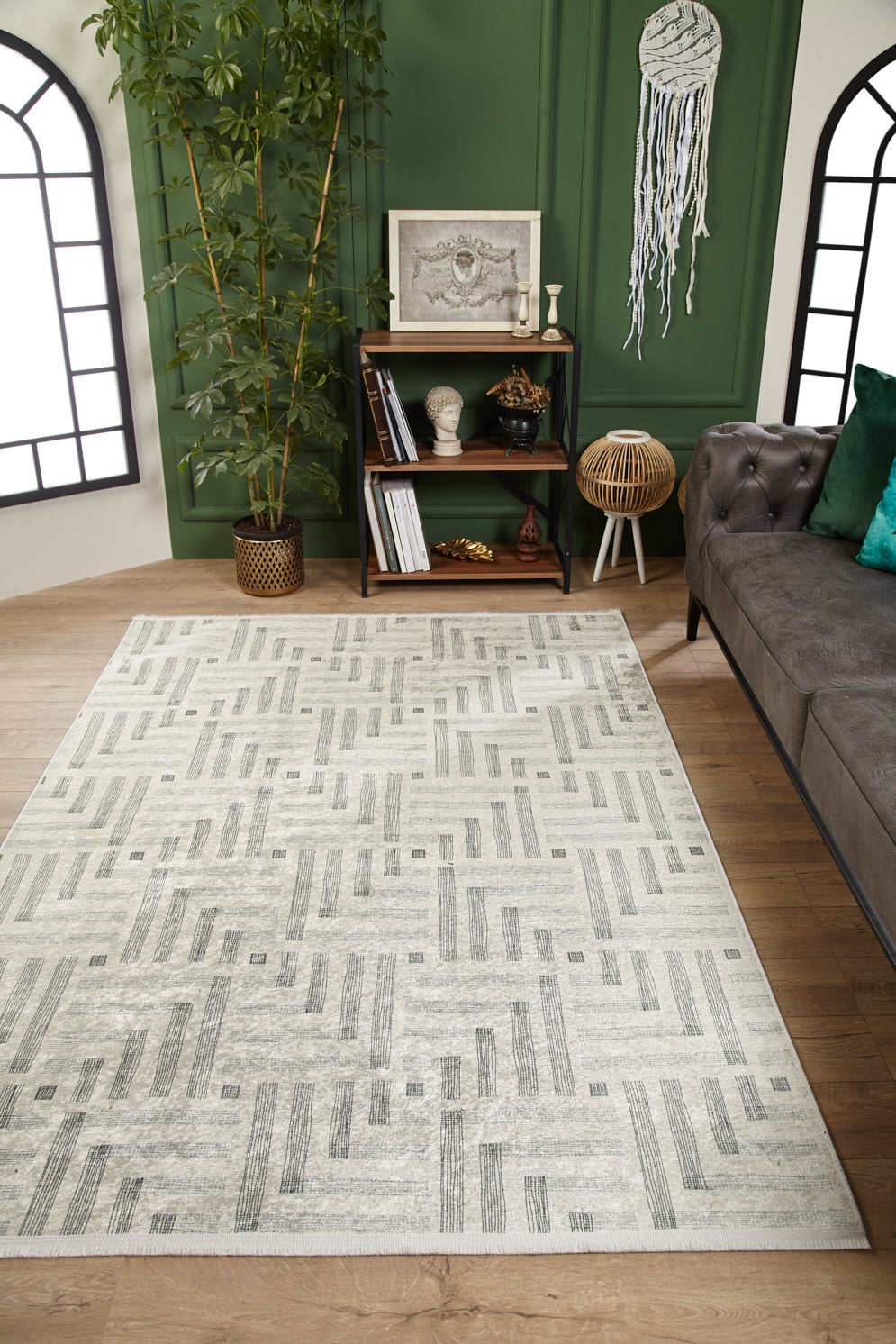 A modern living room featuring the Art Deco Chic - Washable Rug - JR1832 in cream and beige on wooden floors, a gray sofa with green cushions, a decorated green wall, potted plant, bookshelf, and a large window with black trim.