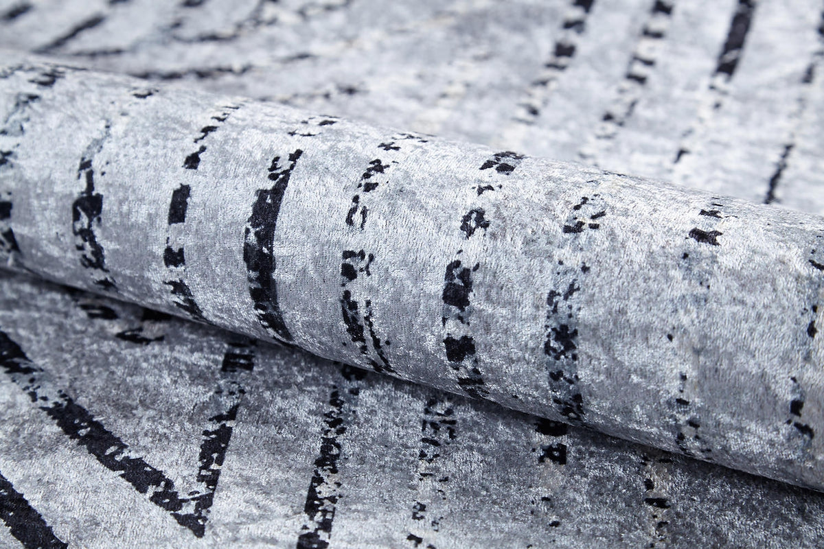 A close-up of the Art Deco Empress - Washable Rug - JR1867 in gray-anthracite, featuring black, irregular vertical lines and a soft, velvety surface. Rolled to show its flexible, plush texture—ideal for high-traffic areas.