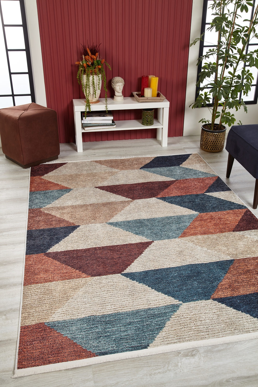 A modern living room featuring the Asymmetrical Wonder - Washable Rug - JR1725 in blue, rust, and beige—ideal for high-traffic spaces—paired with light wood flooring, a white table, decor items, a plant, and a red accent wall with black-trim windows.