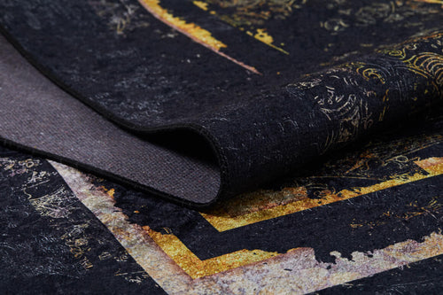 Load image 1 in gallery view. A close-up of the Azure Glow - Washable Rug - JR1337 shows its dark, textured black surface with gold and yellow abstract patterns, slightly folded to reveal layered colors—perfect for high-traffic areas.