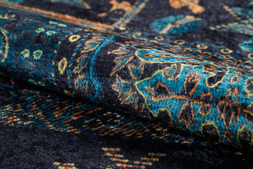 Load image 1 in gallery view. Close-up of the Blossoming Bazaar - Washable Rug (JR1862): a rolled-up, dark-colored rug featuring intricate blue, teal, and yellow floral patterns on black. Its visible texture and durable weave make it ideal for high-traffic areas.