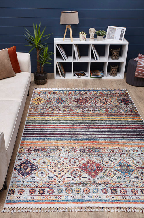 Load image 1 in gallery view. The Bohemian Bouquet - Washable Rug - JR5090, featuring multicolor geometric designs, adorns a wooden floor in a cozy living room with a light sofa, small plant, lamp, and white cube shelf filled with books and decor.