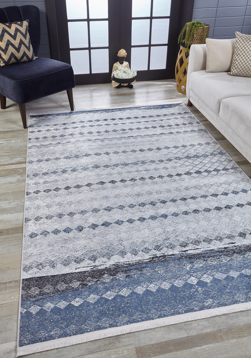 A modern living room features the Bohemian Reverie - Washable Rug - JR1974 (Outlet) US in blue and white on wood floors, paired with a navy chair, chevron pillow, cream sofa, plant stand, and double glass doors for natural light.