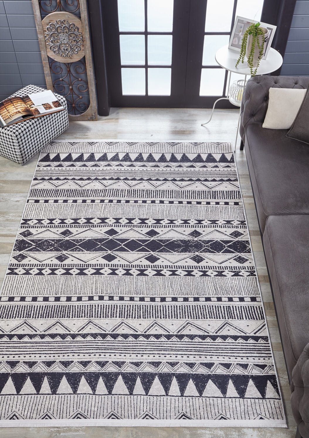 A modern living room with the Bohemian Sunrise - Washable Rug - JR1713 in gray-anthracite, featuring a black-and-white geometric pattern, gray sofa, side table with plant, wall art, and pouf—perfect for high-traffic areas.