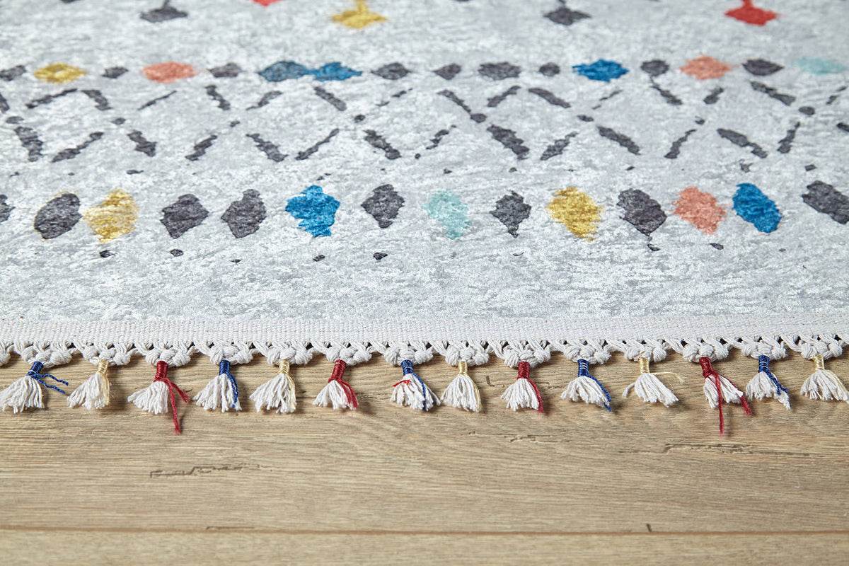 A close-up of the Boho Breeze - Machine-Washable Rug (JR5011) shows its flat weave with tassels, featuring gray, yellow, blue, and orange geometric patterns on a light background atop a wooden floor.