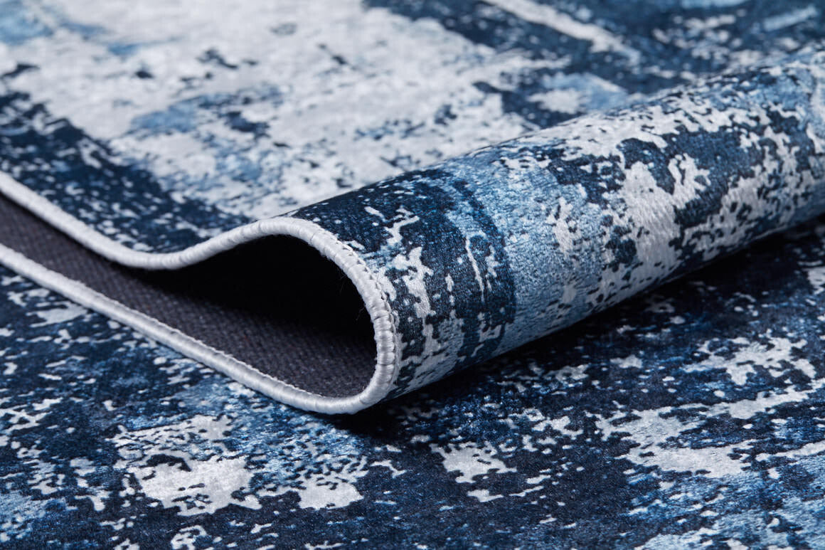 A close-up of the Bold Blooms - Washable Rug - JR836 in blue, featuring an abstract pattern and a folded corner showing a dark underside and stitched edge—ideal for high-traffic spaces.