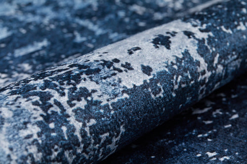 Load image 1 in gallery view. A close-up of the Bold Blooms - Washable Rug - JR836, featuring a blue abstract marbled pattern in black and white, slightly rolled up. This washable rug is ideal for high-traffic areas, offering both style and practicality.