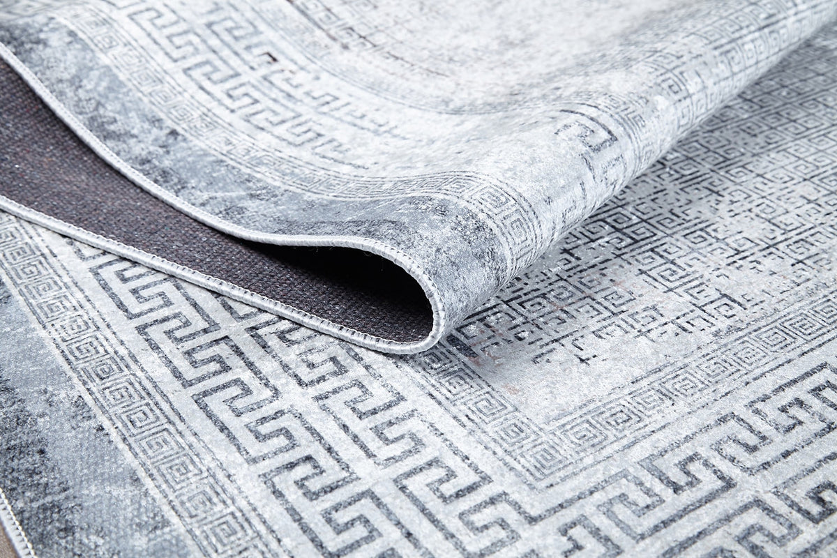 A close-up of the Bold Borderline - Washable Rug - JR1984 in gray-anthracite, featuring a Greek key pattern and distressed texture, with one edge folded to reveal its washable underside—ideal for high-traffic areas.