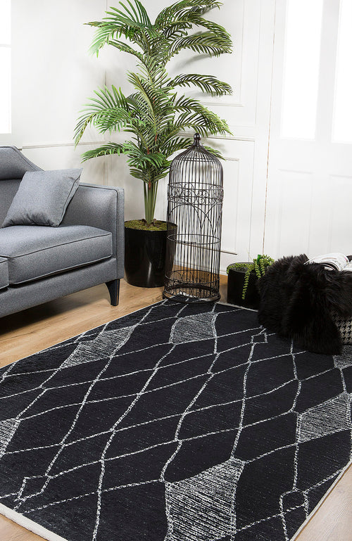 Load image 1 in gallery view. A modern living room features a gray sofa, a large leafy potted plant, black decorative birdcage, black pillows on a light wood floor, and the Byzantine Beauty - Washable Rug - JR1037—perfect for high-traffic areas with its stylish black and white pattern.