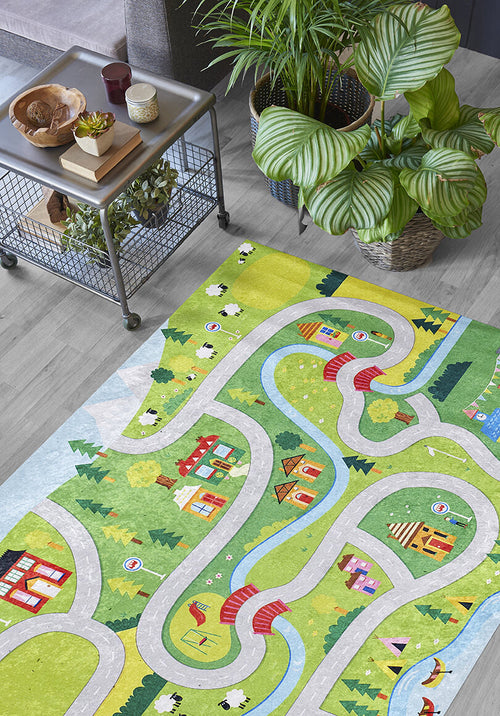 Bild 1 in Galerieansicht laden. The City Adventure - Machine-Washable Rug JRC039, featuring colorful winding roads, houses, rivers, and trees, is spread on a wooden floor beside a metal side table and green potted plants—cheerful, child-friendly, and easy to clean.