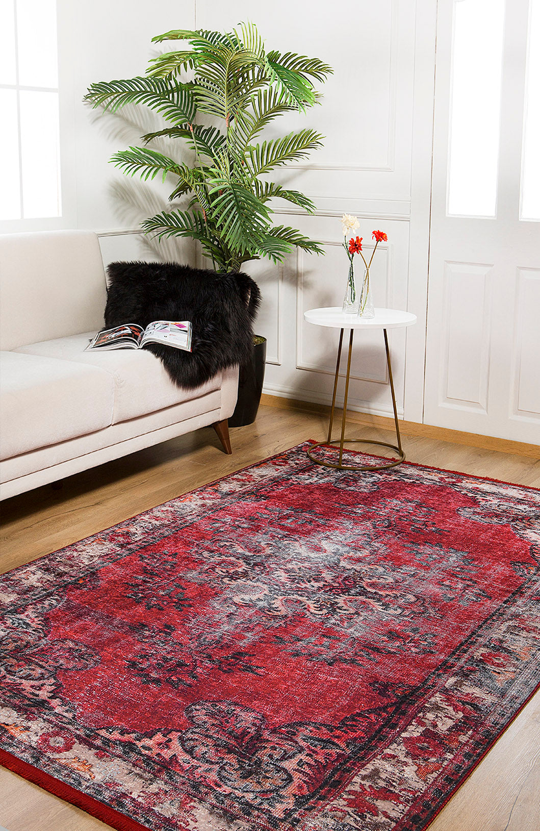 A cozy living room features the Classic Revival - Washable Rug - JR1374 in red, a cream sofa with a black pillow and magazine, a white round side table with flowers, and a potted plant by large white windows—ideal for high-traffic spaces.