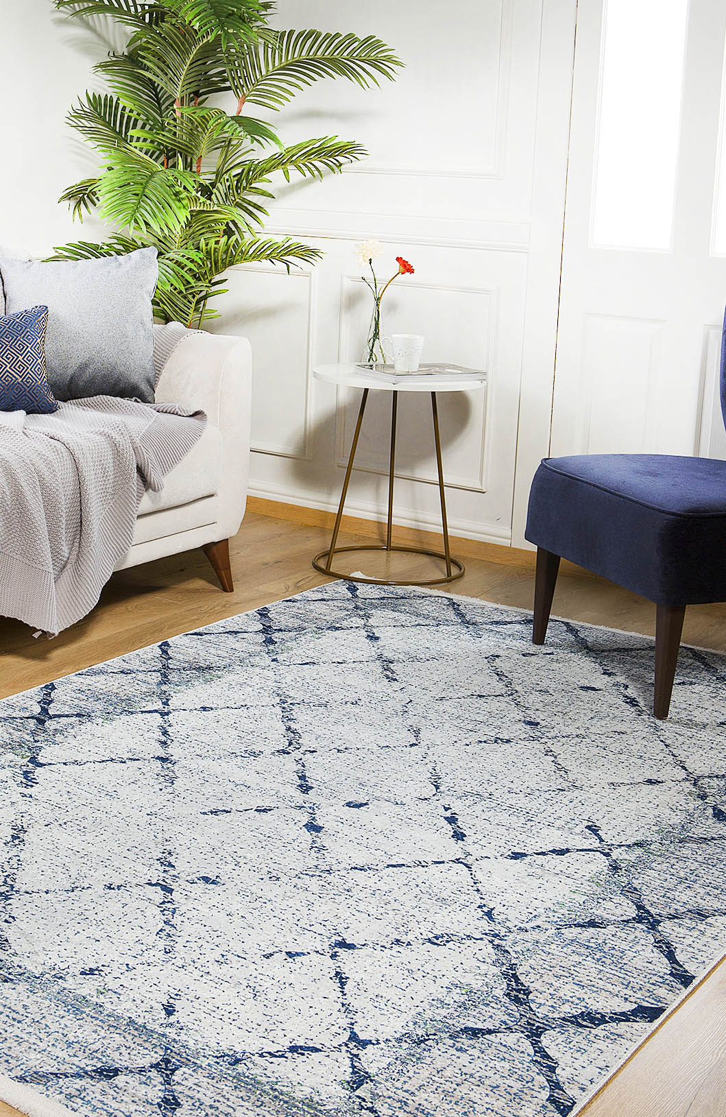A cozy living room with a white sofa, blue cushioned chair, leafy plant, small round side table with flowers, and the Classic Trellis - Washable Rug - JR710 on a wooden floor, illuminated by sunlight streaming through a window.