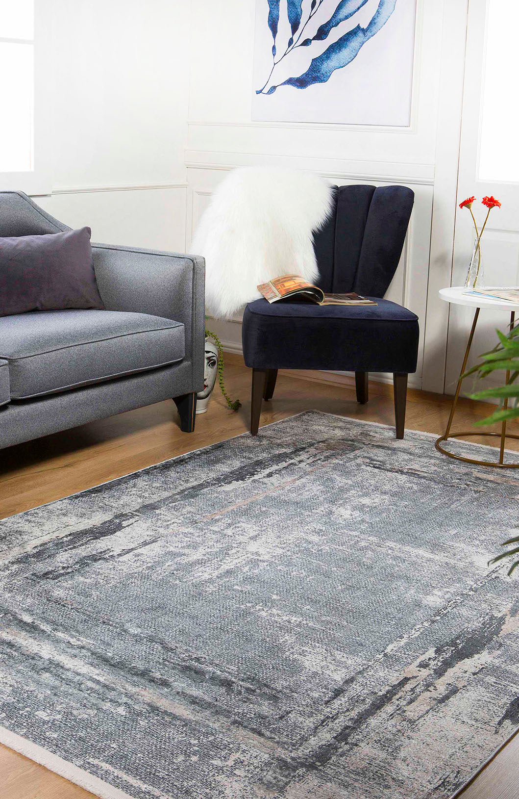 A modern living room features a gray sofa, dark blue chair, and the Classic and Bold - Washable Rug - JR1529—ideal for high-traffic areas. A small side table with red flowers, white throw, wall art, and a face sculpture complete the look.