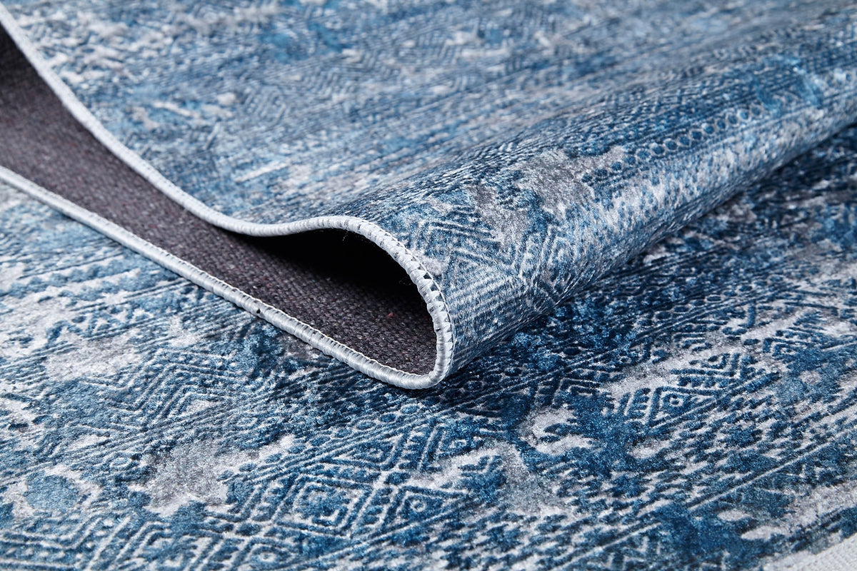 Close-up of the Contemporary Serenity - Machine-Washable Rug JR1884 in blue and gray, featuring intricate geometric and floral patterns with a lifted corner revealing its dark underside—durable and ideal for high-traffic areas.