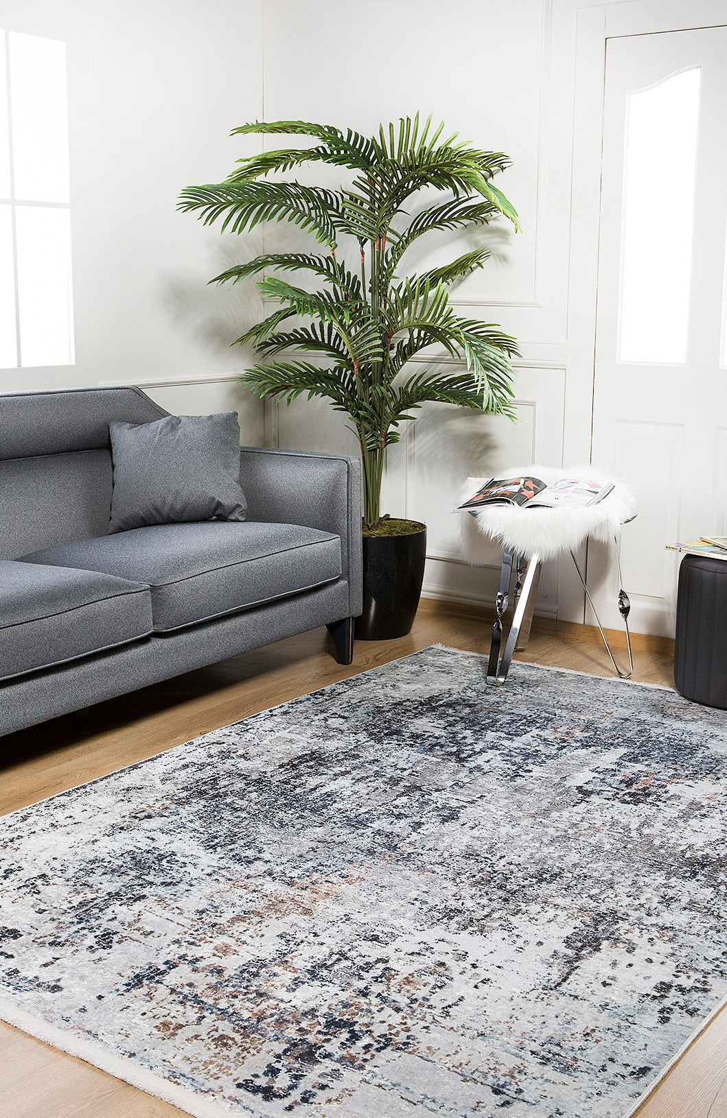 Modern living room featuring the gray sofa and a Creative Currents - Washable Rug - JR835, ideal for high-traffic areas. Natural light highlights the palm plant, crisp white walls, and a small table with magazines—perfect for everyday living.