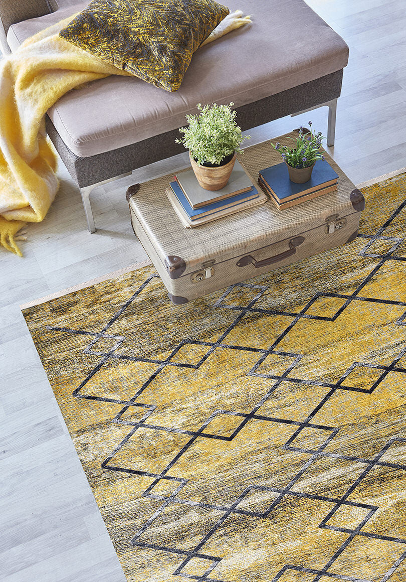 A living room with the yellow-gold Crossed Connections Washable Rug (JR1181) for high-traffic areas, a beige ottoman with a yellow throw and pillow, plus a vintage suitcase coffee table displaying books, a plant, and a tray.