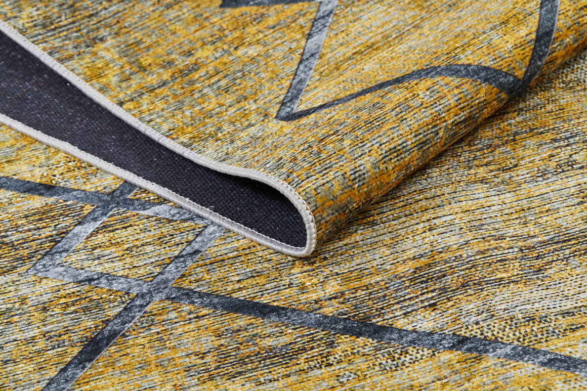 A close-up of the Crossed Connections - Washable Rug - JR1181 in yellow-gold with a gray geometric pattern, featuring one corner lifted to reveal its dark underside—ideal for high-traffic areas.