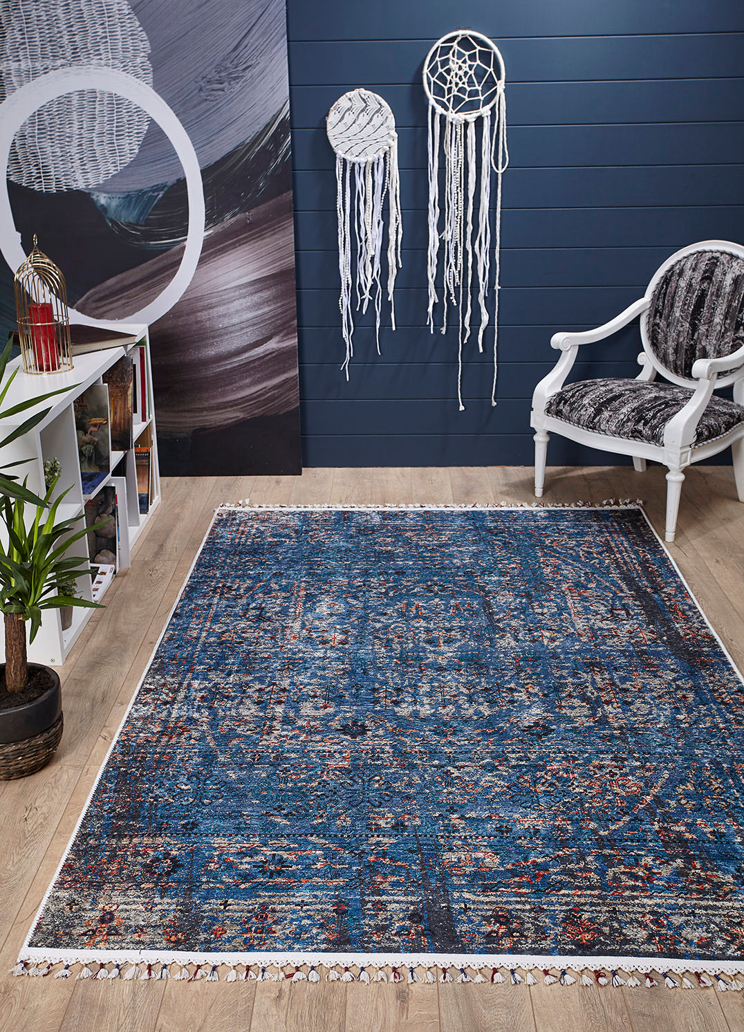 A modern living room features the Desert Diamond - Washable Rug - JR5071 in blue pattern for high-traffic areas, a white fabric armchair, two large dreamcatchers on a dark blue wall, bookshelf, potted plant, and wood flooring.
