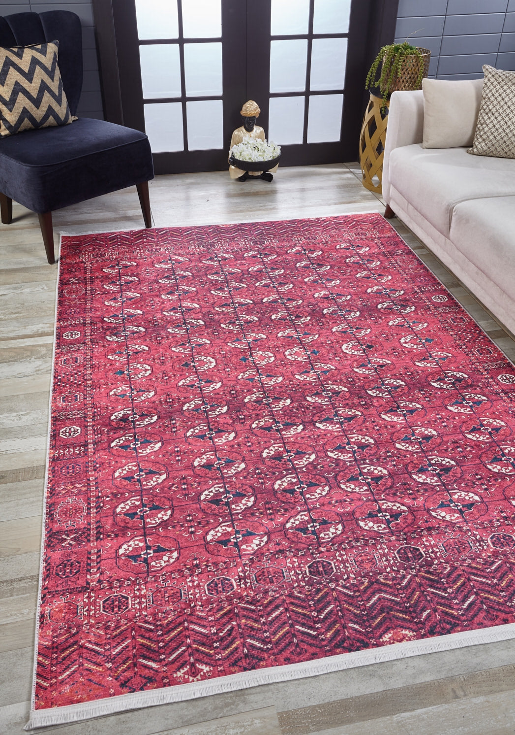 A living room features the Desert Dreamscape - Washable Rug - JR1940 in red patterns on light wood flooring, paired with a dark blue chair, chevron pillow, white sofa, and a vase by a frosted glass window.