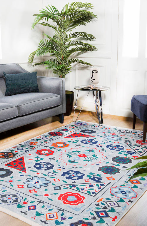 Load image 1 in gallery view. A modern living room with a gray sofa, blue ottoman, glass side table with a face vase, large green plant, and the Desert Drift - Washable Rug - JR1523—ideal for high-traffic areas—on light wood flooring.