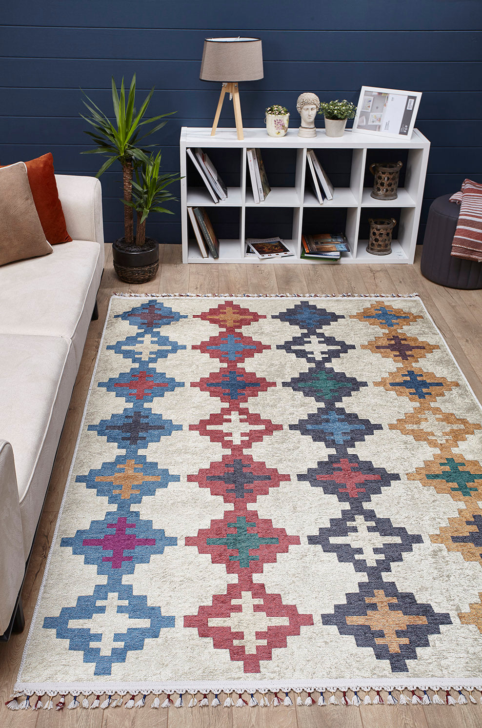 A living room features a beige sofa, patterned cushions, a Desert Mirage - Washable Rug - JR5033 with colorful geometric design, a white shelf holding books and decor, a potted plant, and a table lamp set against dark blue walls and wooden flooring.