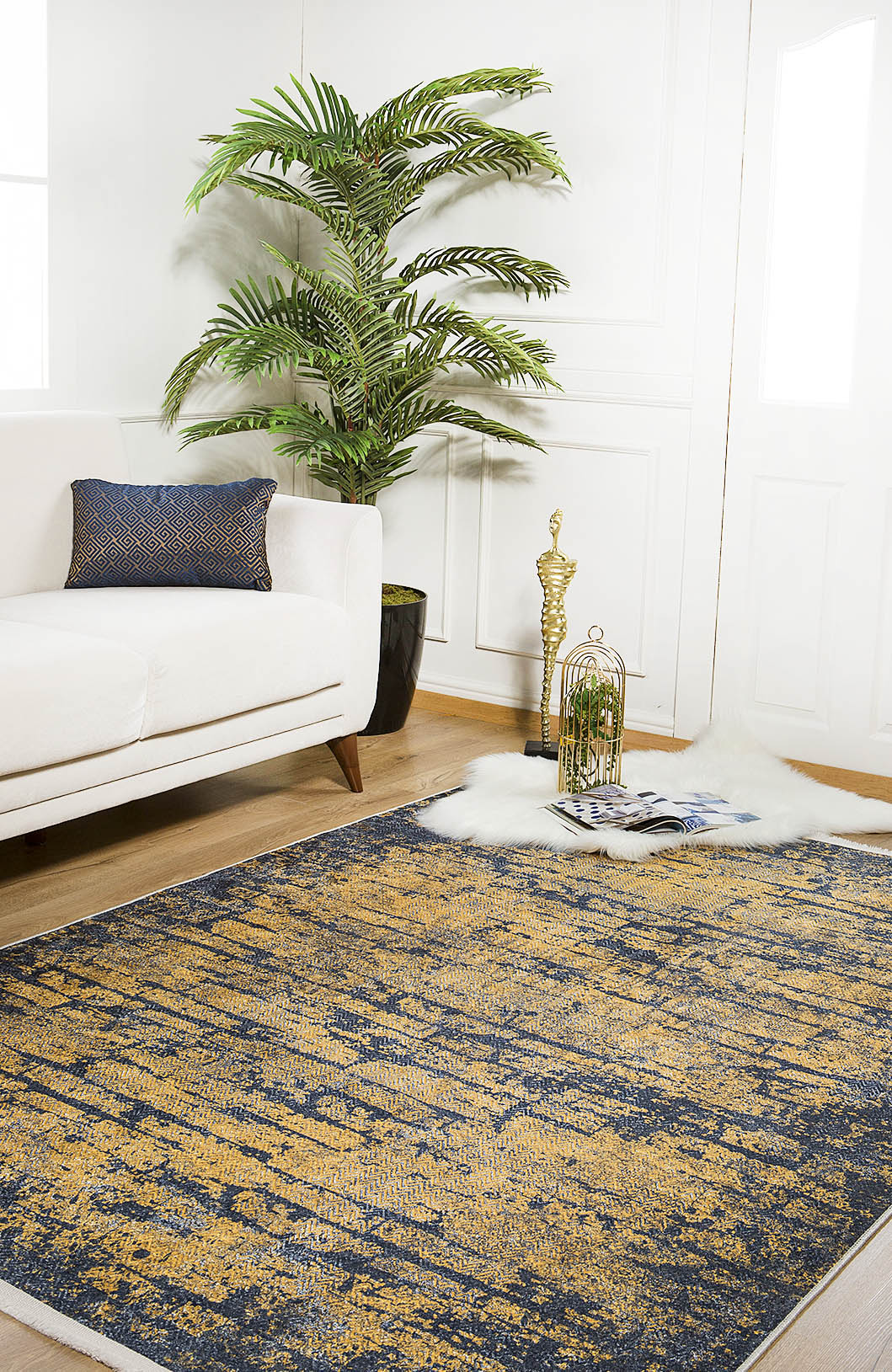 A modern living room featuring a white sofa, the Diagonal Dash - Washable Rug - JR1310 in blue and gold for high-traffic areas, a green potted plant, a decorative gold sculpture, magazines, and a fluffy white rug on wooden floors.