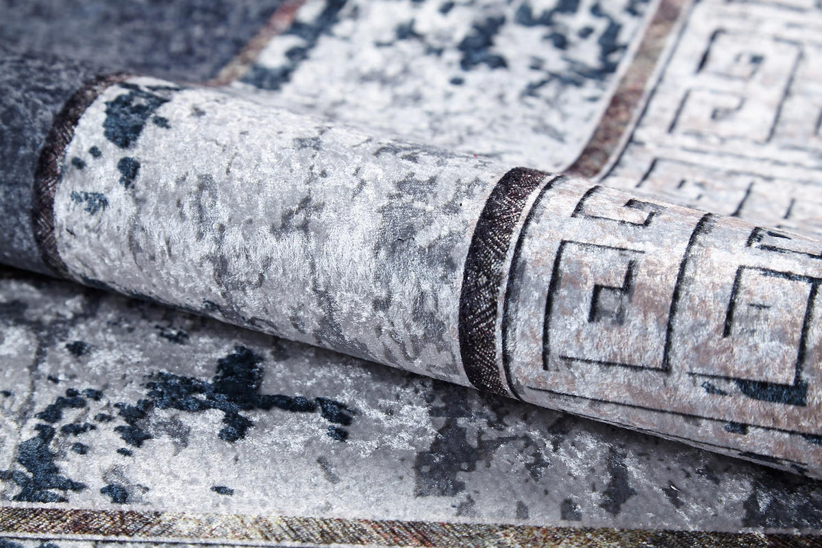 A close-up of the Dramatic Boundaries - Washable Rug (JR1810), folded to show its gray, textured, mottled pattern and geometric border in dark and light tones—perfect for high-traffic areas.