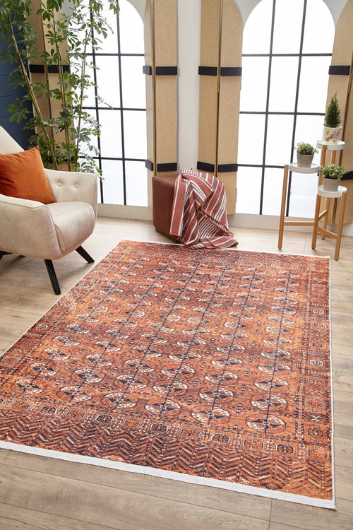 Load image 1 in gallery view. A living room featuring the Earthy Enigma - Washable Rug - JR1937 in orange, ideal for high-traffic areas, a white armchair with orange pillows, pouf with striped throw, plant stand, large windows, and tall corner plants.