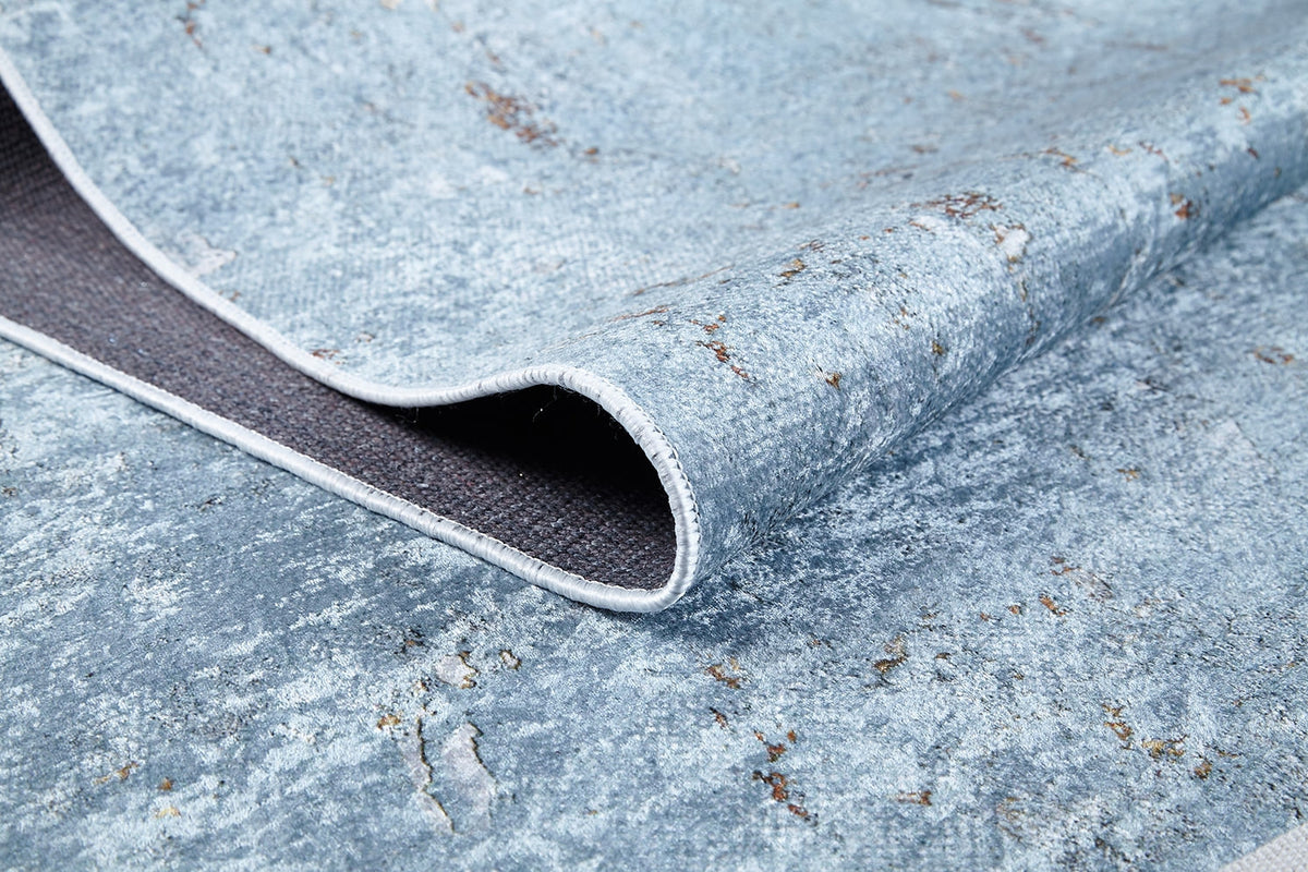 A close-up of the Earthy Sands - Washable Rug - JR1819 shows its blue and grey distressed texture, one edge lifted to reveal the dark underside and stitched border—perfect for high-traffic areas.