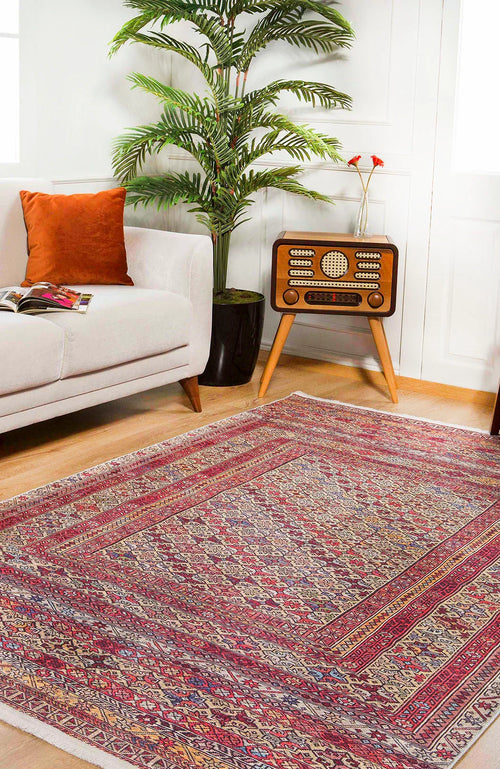 Load image 1 in gallery view. A living room featuring the Elegant Embellishments Washable Rug - JR43 in red for high-traffic areas, a white sofa with an orange cushion, a vintage radio on wooden legs, a tall potted plant, and a magazine on the sofa.