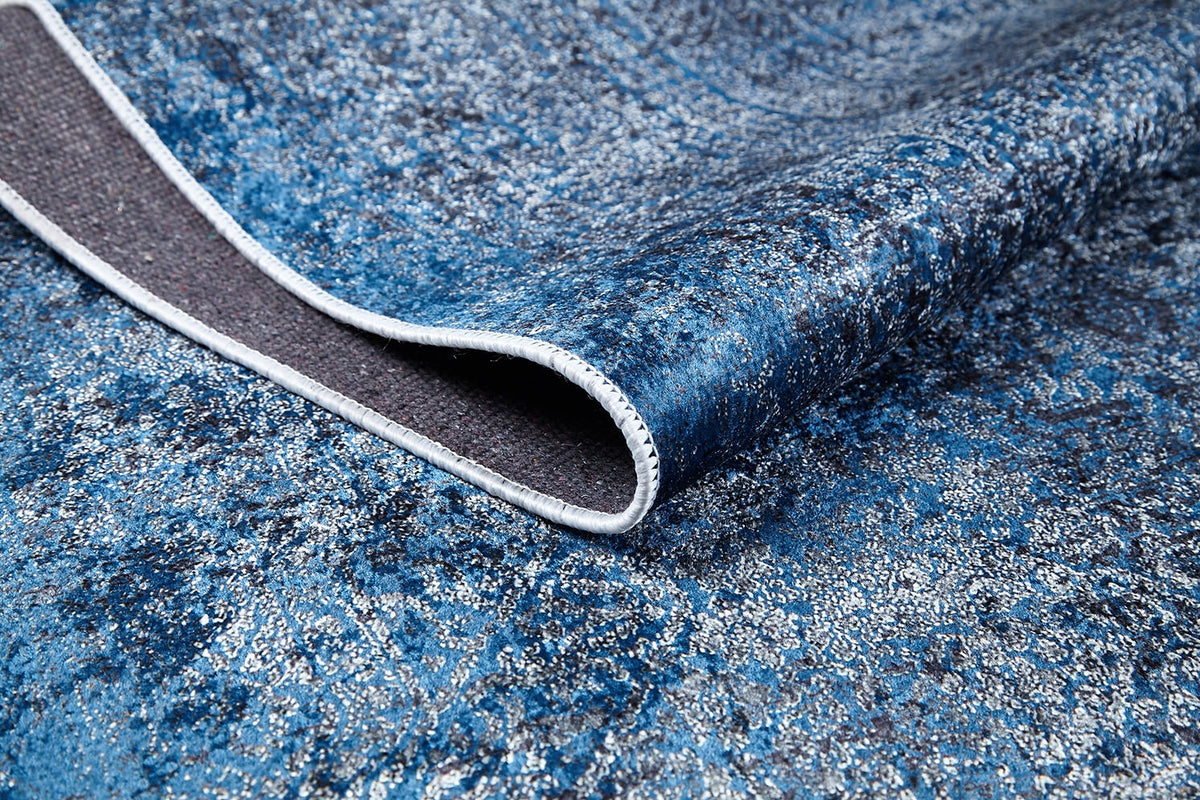A close-up of the Elegant Vine - Washable Rug - JR1847 in blue and silver shows its flat weave texture, with one corner folded to reveal the underside and detailed edge stitching.
