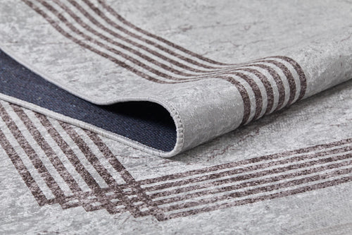 Load image 1 in gallery view. A close-up of the Elegant and Chic - Washable Rug - JR1509 shows its light gray surface with brown border lines and a lifted corner revealing a dark, textured underside—ideal for high-traffic spaces.