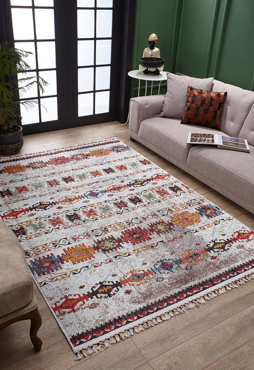 Bild 1 in Galerieansicht laden. The Ethno Expanse Machine-Washable Rug (JR5046), colorful and adorned with tassels, brightens a wooden floor in a modern living room featuring a light gray sofa, decorative pillow, side table, plant, and large windowed doors.