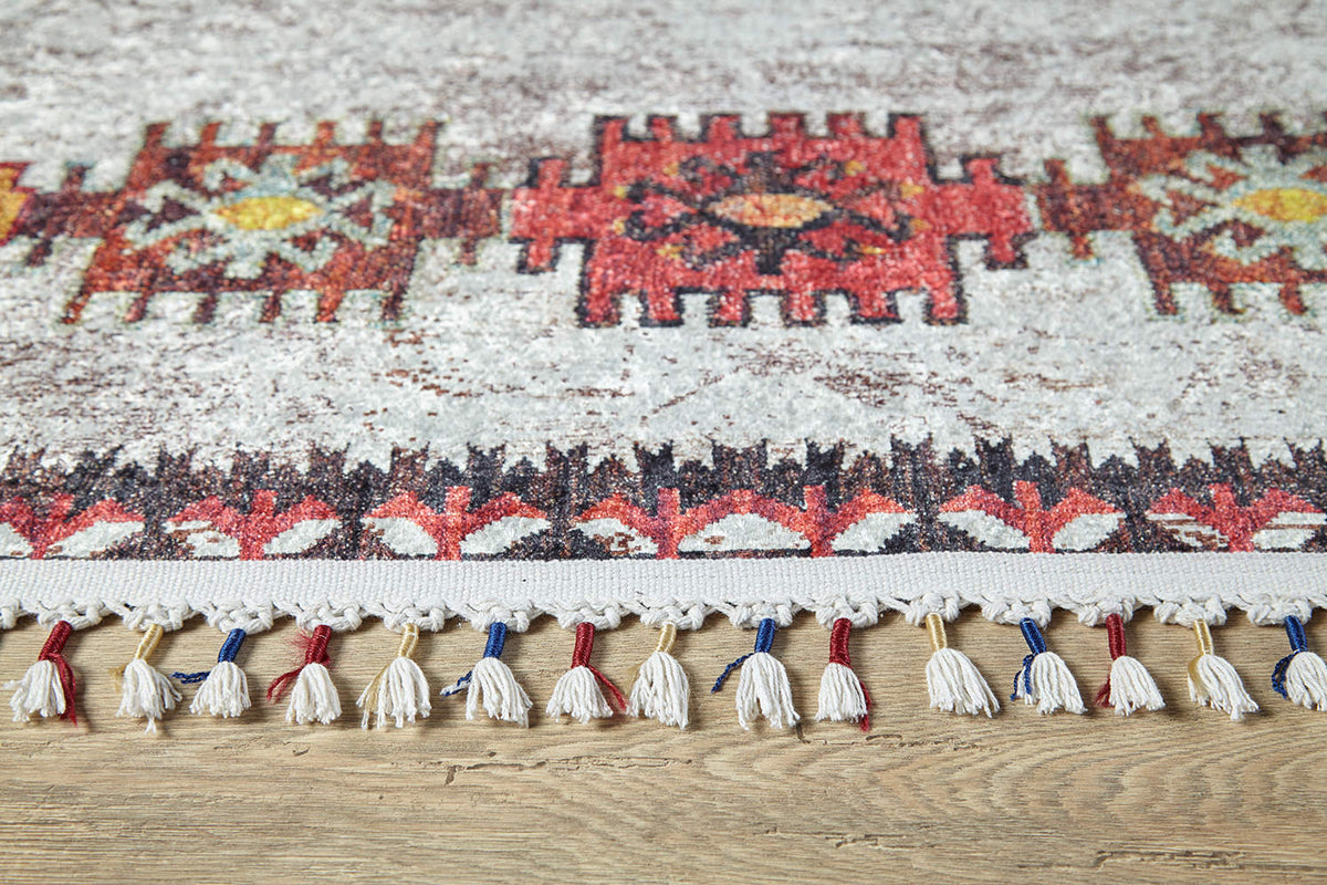 The Ethno Expanse Machine-Washable Rug (JR5046) features red, yellow, and black geometric patterns with white fringes accented by red and blue threads—ideal for high-traffic areas and shown on a wooden floor.