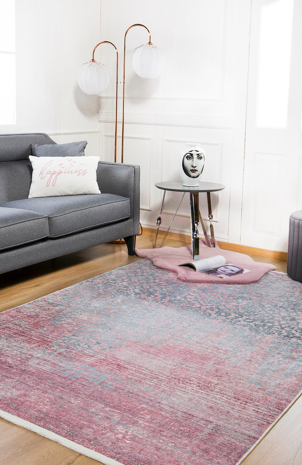 A modern living room with a gray sofa, white happiness pillow, copper floor lamp, round side table with decorative face vase, and the Floral Filigree Washable Rug JR341 in soft pink and gray—perfect for allergy-sensitive homes.