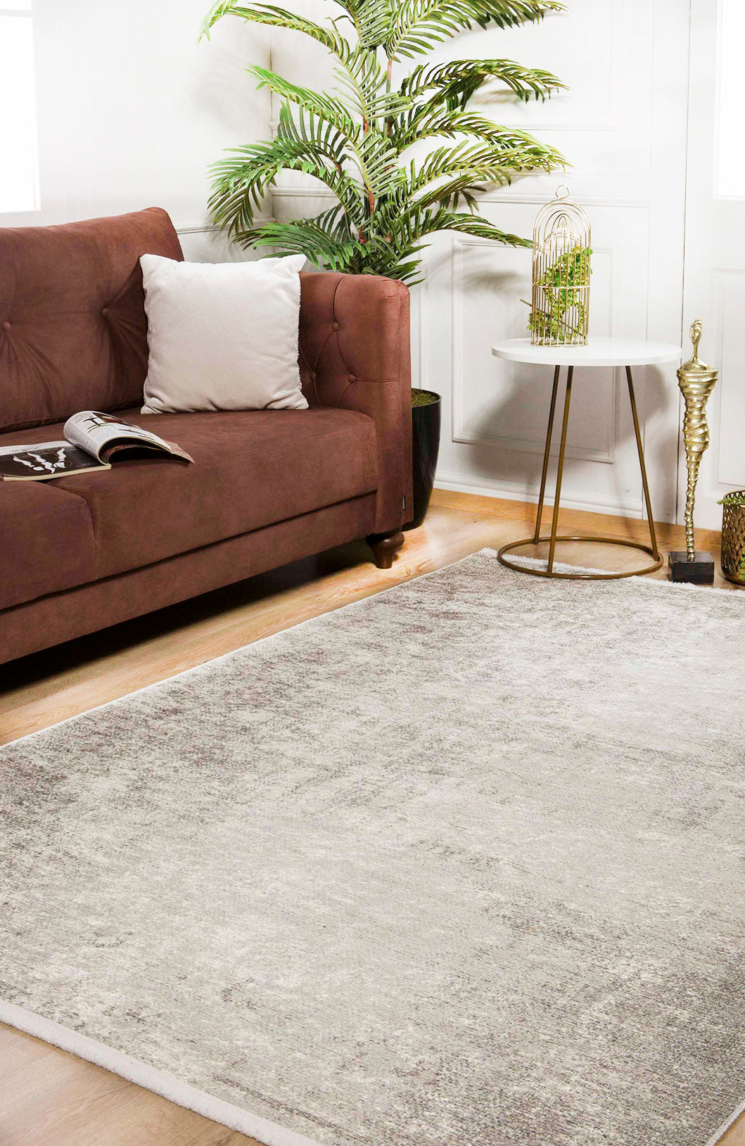 A modern living room featuring a brown sofa, white pillow, open magazine, Forest Canopy - Washable Rug - JR800, round white side table, decorative accents, potted plant, and bright natural light streaming through windows.