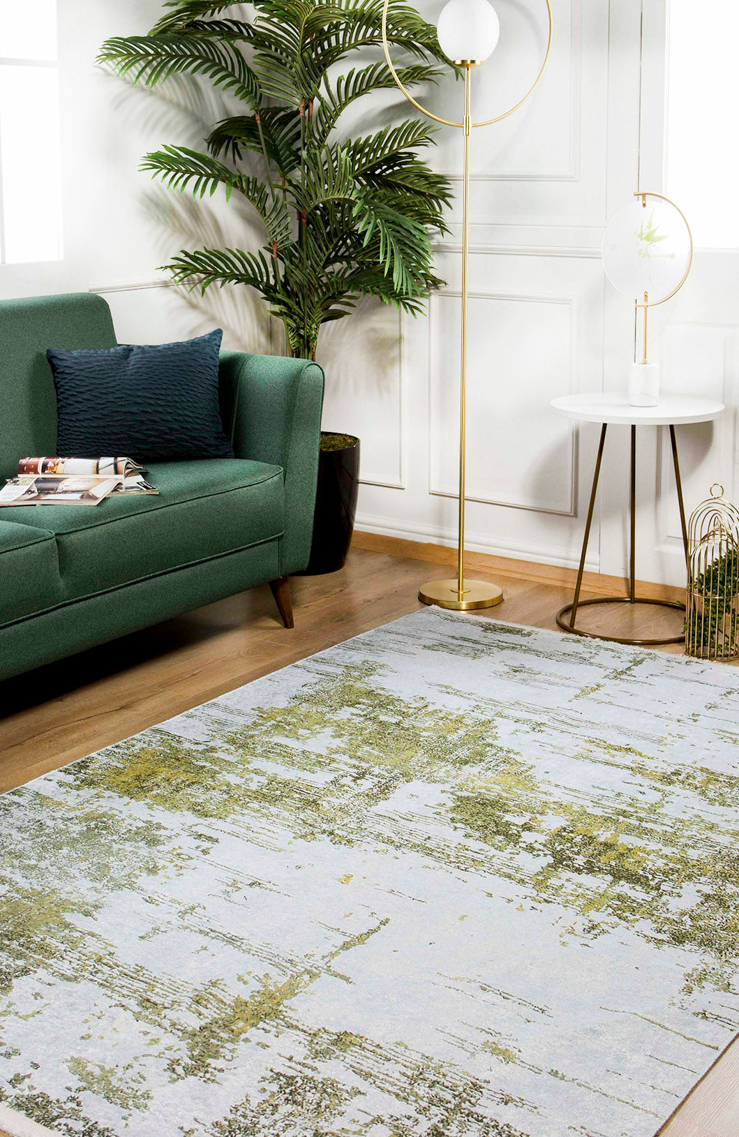 A modern living room with a green sofa, potted plant, gold floor lamp, white side table with a globe lamp, and the allergy-friendly Geometric Geometry - Washable Rug - JR1290 featuring green and gray abstract patterns on wood floors.