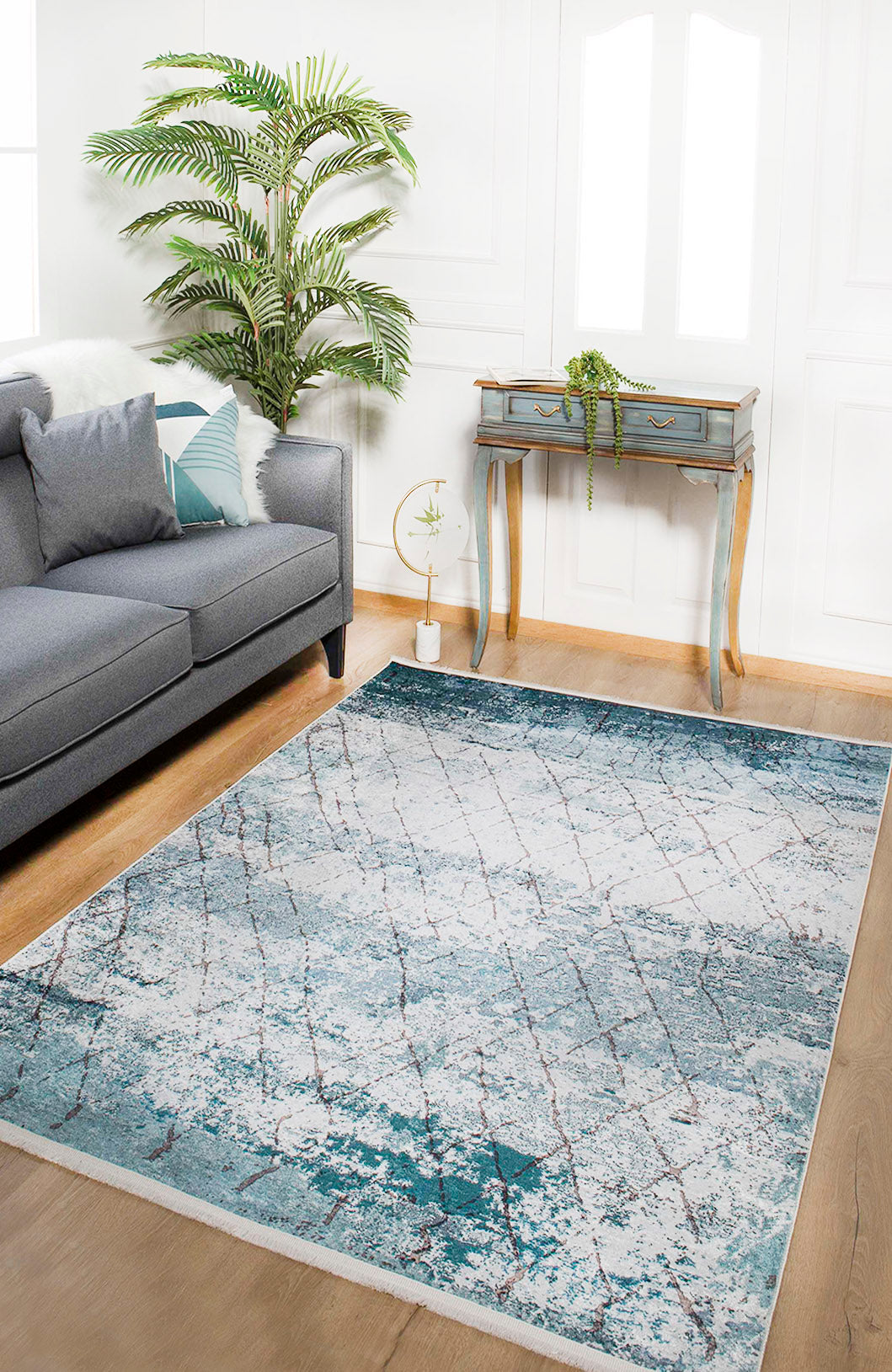 A modern living room with a gray sofa, teal and white pillows, the Geometric Glimmers - Washable Rug - JR1423 ideal for high-traffic areas, a small desk with a plant, a potted palm, and plenty of natural light from the window.