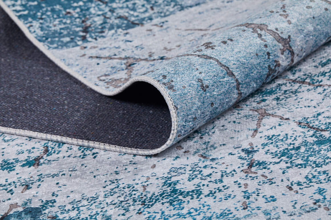 A close-up of the Geometric Glimmers - Washable Rug - JR1423 shows its blue and gray marbled pattern, a folded corner revealing the dark underside and stitched edges—ideal for high-traffic areas.