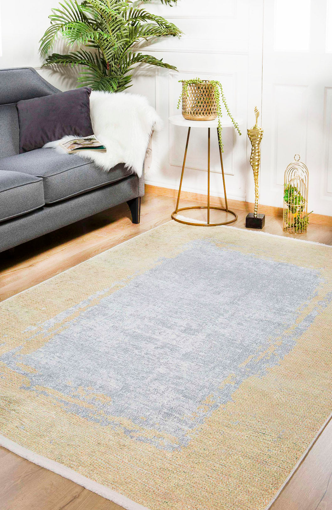 A modern living room with a grey sofa, the Geometric Trellis - Washable Rug - JR931 in soft grey and beige, a gold side table with a potted plant, a gold floor sculpture, a birdcage, and a tall leafy plant in the corner.