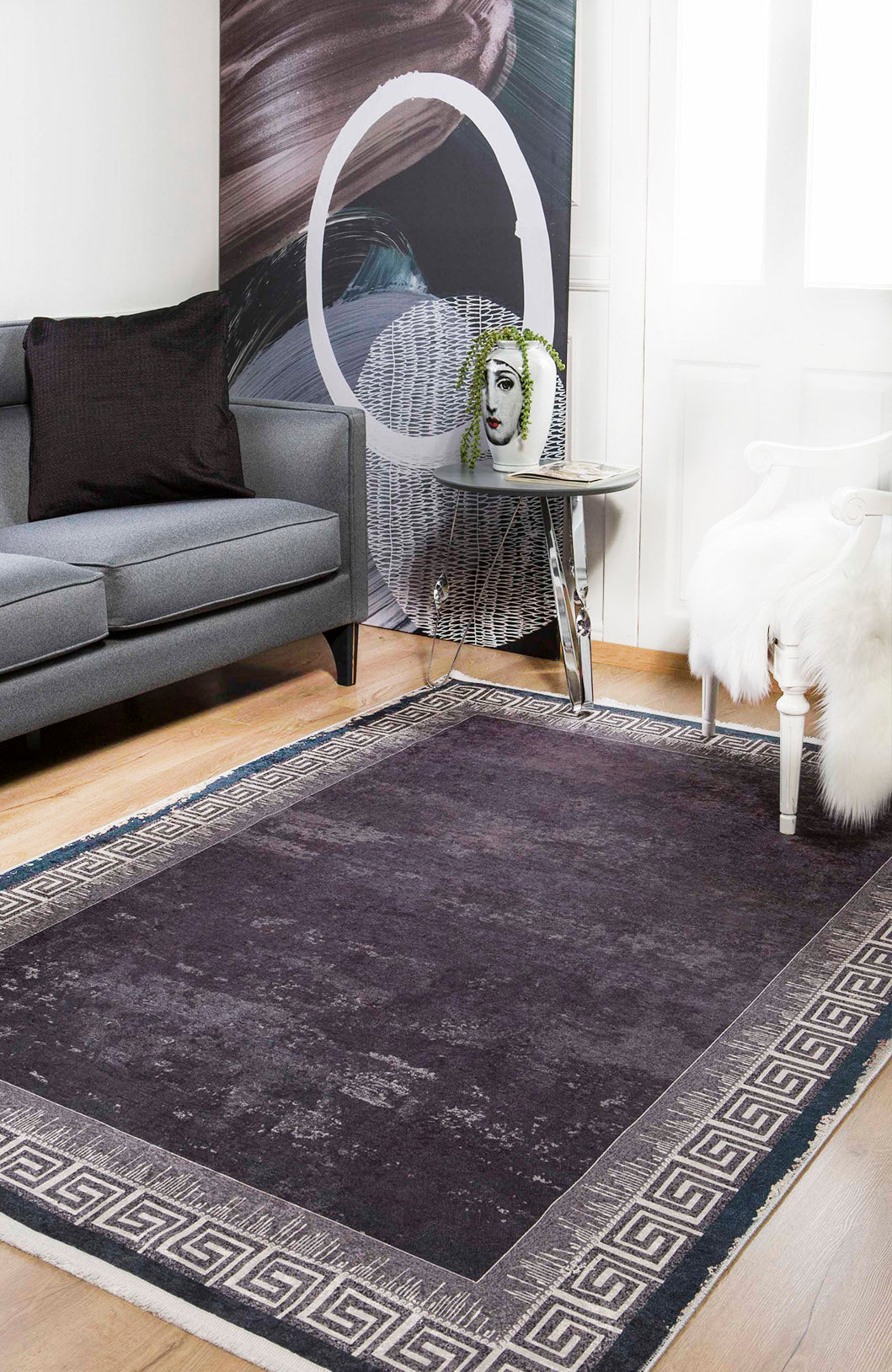 A modern living room with the Geometric and Chic - Washable Rug - JR1273 in dark gray with a Greek key border, a gray sofa, black pillow, white chair with fur throw, face vase with greenery on a table, and abstract wall art. Great for high-traffic areas.