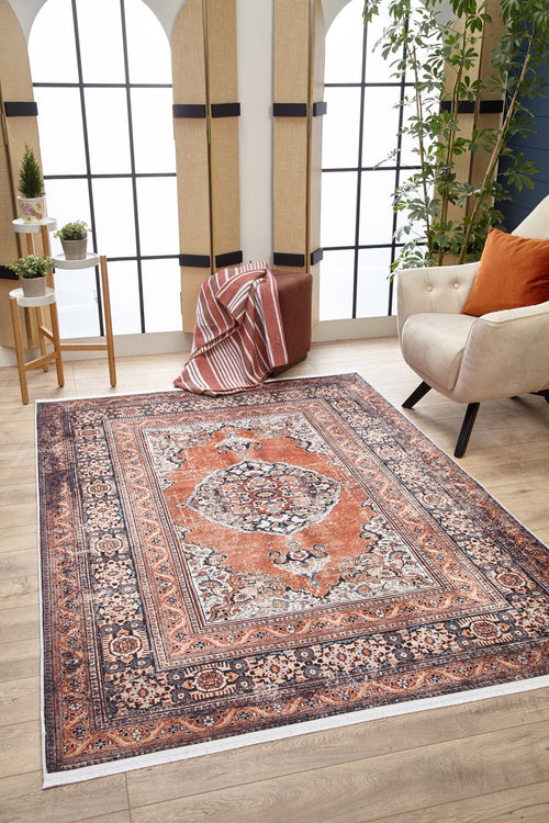 Load image 1 in gallery view. A cozy living room with the Golden Legacy - Washable Rug - JR1931 in orange and navy, ideal for high-traffic areas, a cream armchair with rust cushion, a small round table with plants, a pouf draped with a striped blanket, and large arched windows.