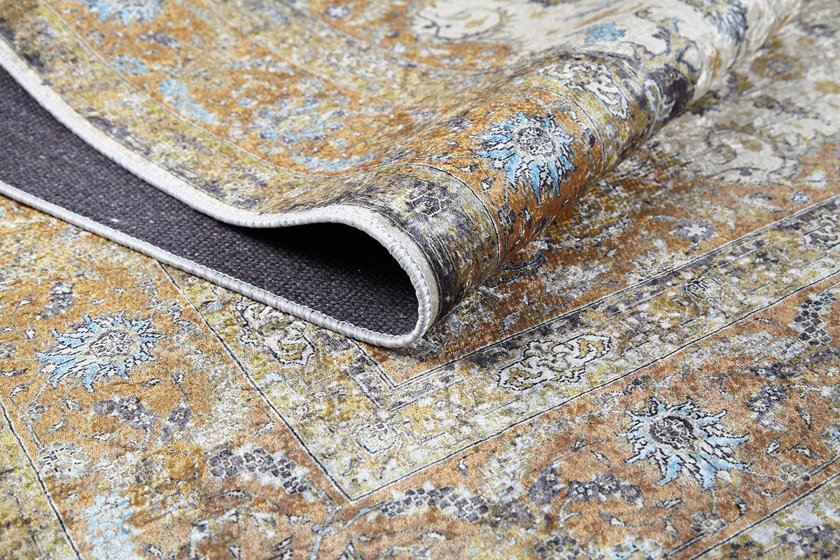 A close-up of the Golden Oasis - Washable Rug - JR1930 shows a folded corner revealing its textured backing. The rug displays an intricate yellow, blue, and white design with a slightly distressed look, ideal for high-traffic areas.