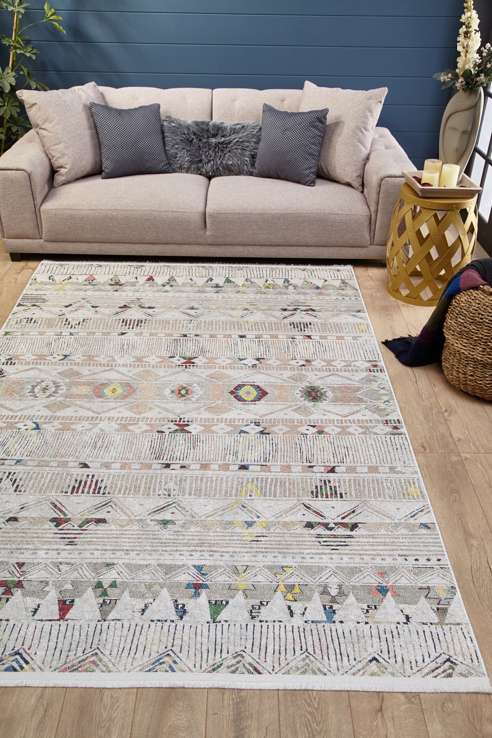 A beige sofa with gray and fur pillows sits against a blue wall. The Gypsy Glam - Washable Rug - JR1716, featuring colorful geometric designs, covers the wooden floor. A side table with candles and a woven basket are nearby.