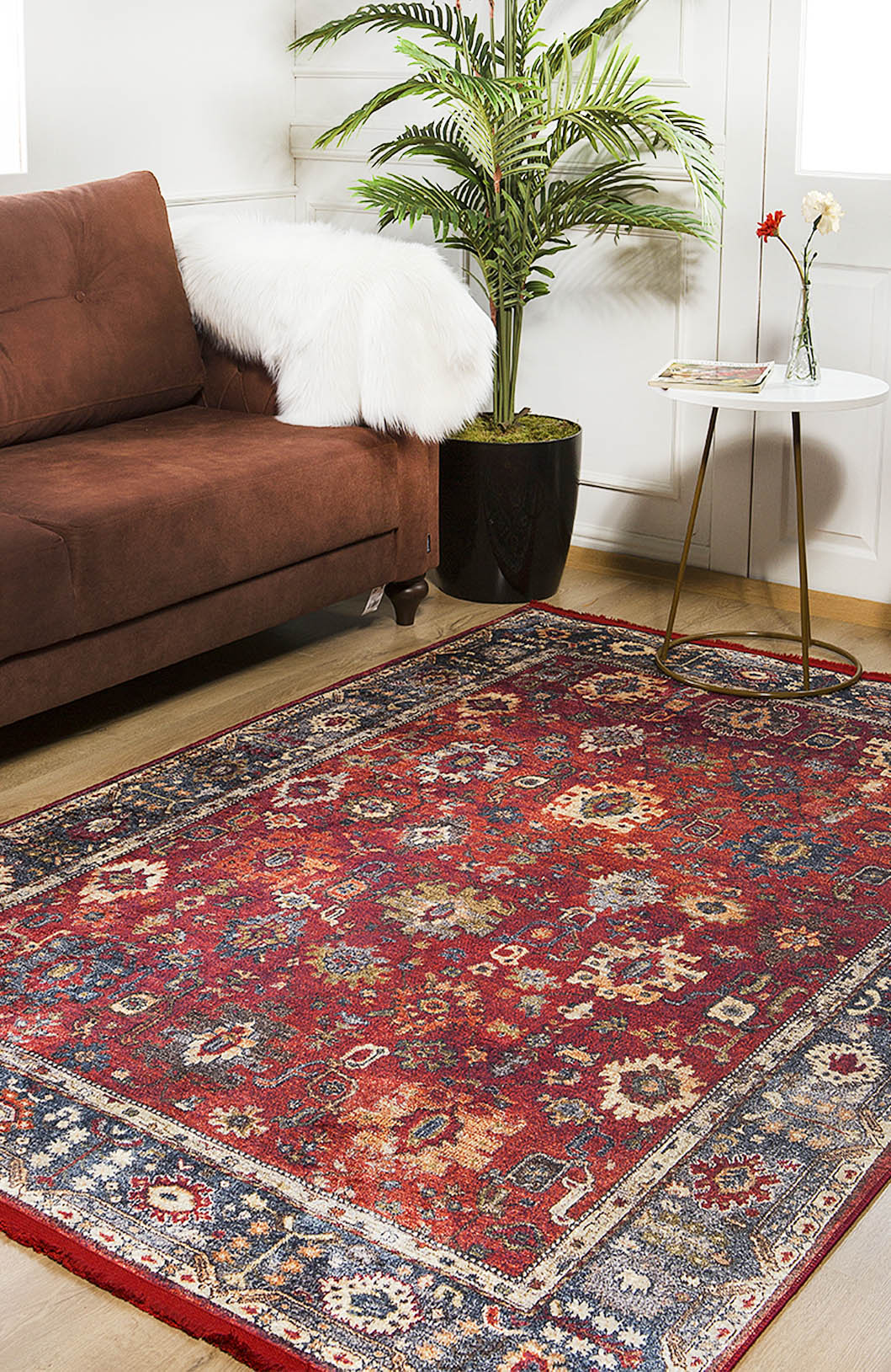 A living room with a brown sofa, the Historic Hues - Washable Rug - JR898 (Outlet) US in red for high-traffic areas, a round white side table topped with red and white flowers, a book, and a potted plant by the window.