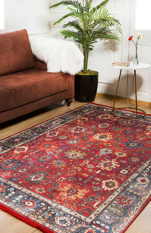 Load image 1 in gallery view. A living room with a brown sofa, the Historic Hues - Washable Rug - JR898 (Outlet) US in red for high-traffic areas, a round white side table topped with red and white flowers, a book, and a potted plant by the window.