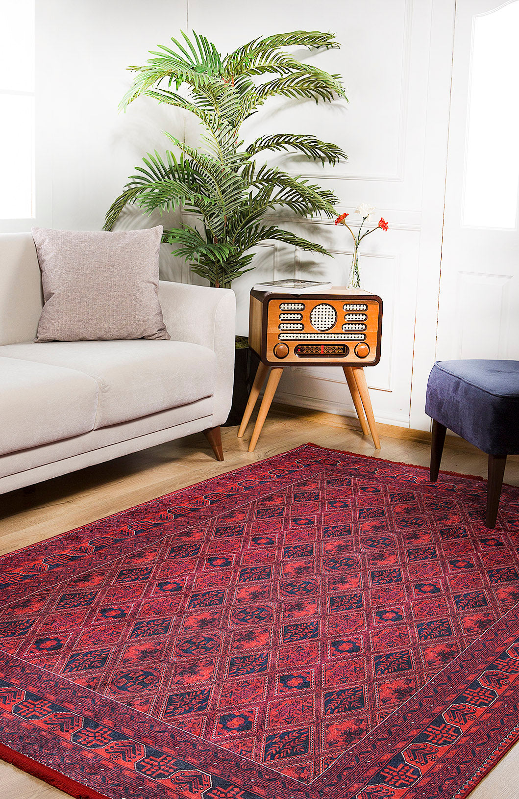A cozy living room features the Imperial Impressions - Washable Rug - JR1132, ideal for high-traffic areas, a white sofa with a gray pillow, vintage radio on a wooden stand, potted plant, dark blue ottoman, and bright natural light.