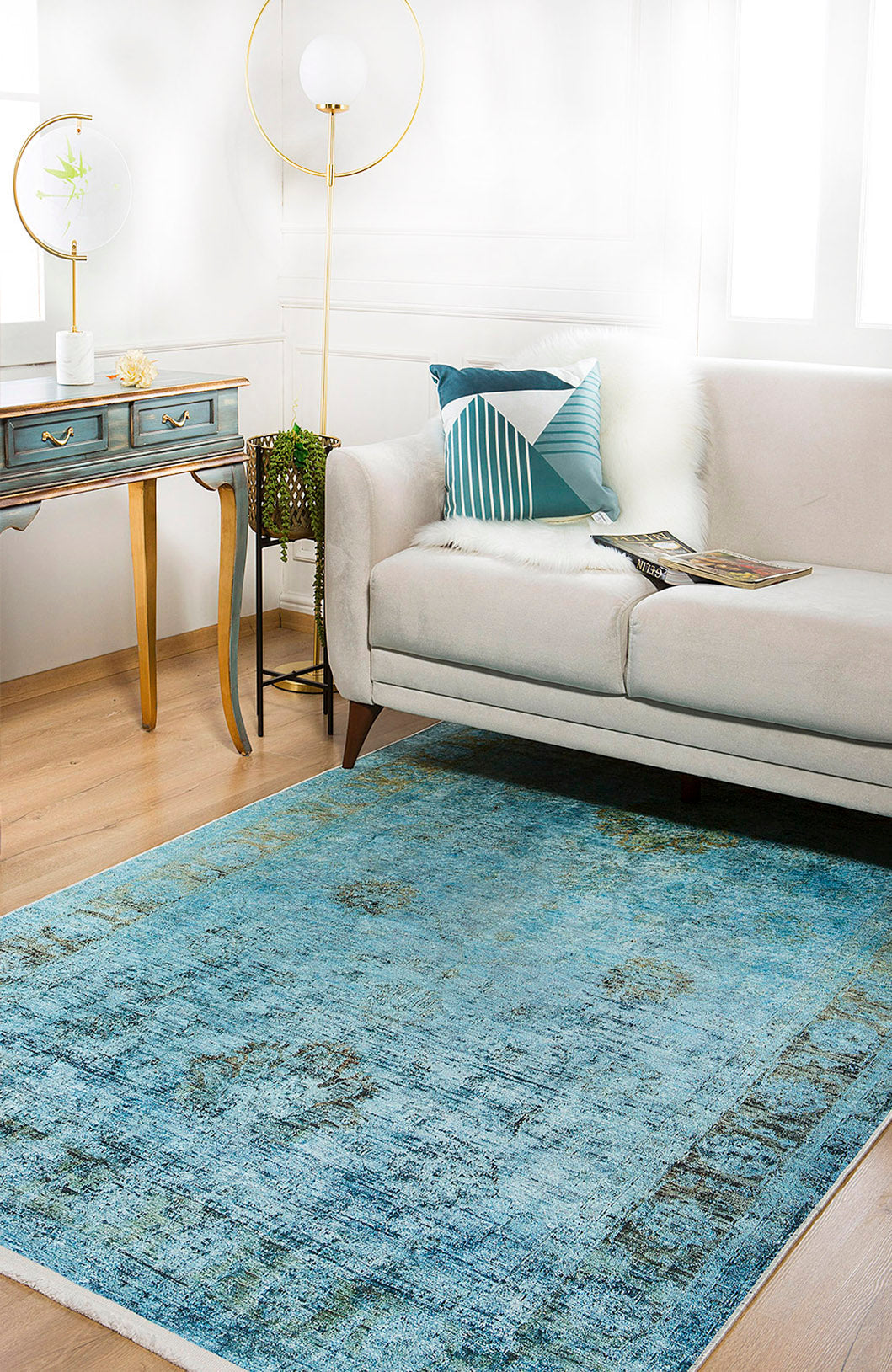 A modern living room with a light gray sofa, geometric blue pillow, the Imperial Jewel - Washable Rug - JR162 (blue with gold accents) ideal for high-traffic areas, a small blue desk with gold legs, round lamp, and potted plant on wooden flooring.