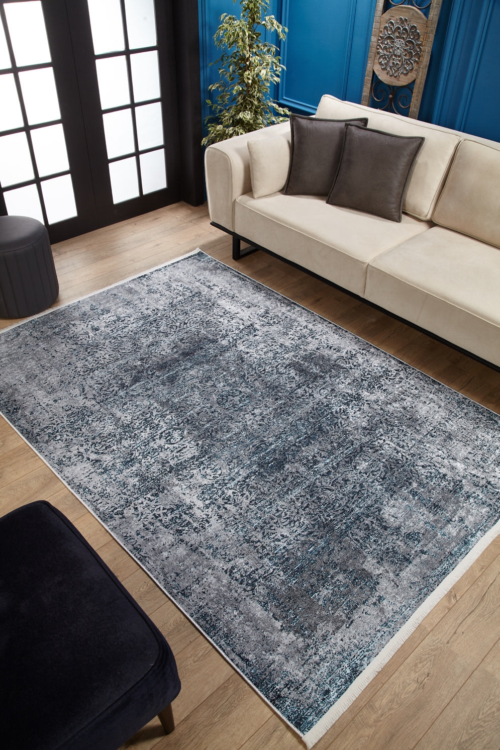 A modern living room with a beige sofa, dark accent pillows, the Imperial Lace Washable Rug (JR1607) in blue on wooden floors—ideal for busy spaces—a black ottoman, green plant, and blue paneled walls with decorative art.